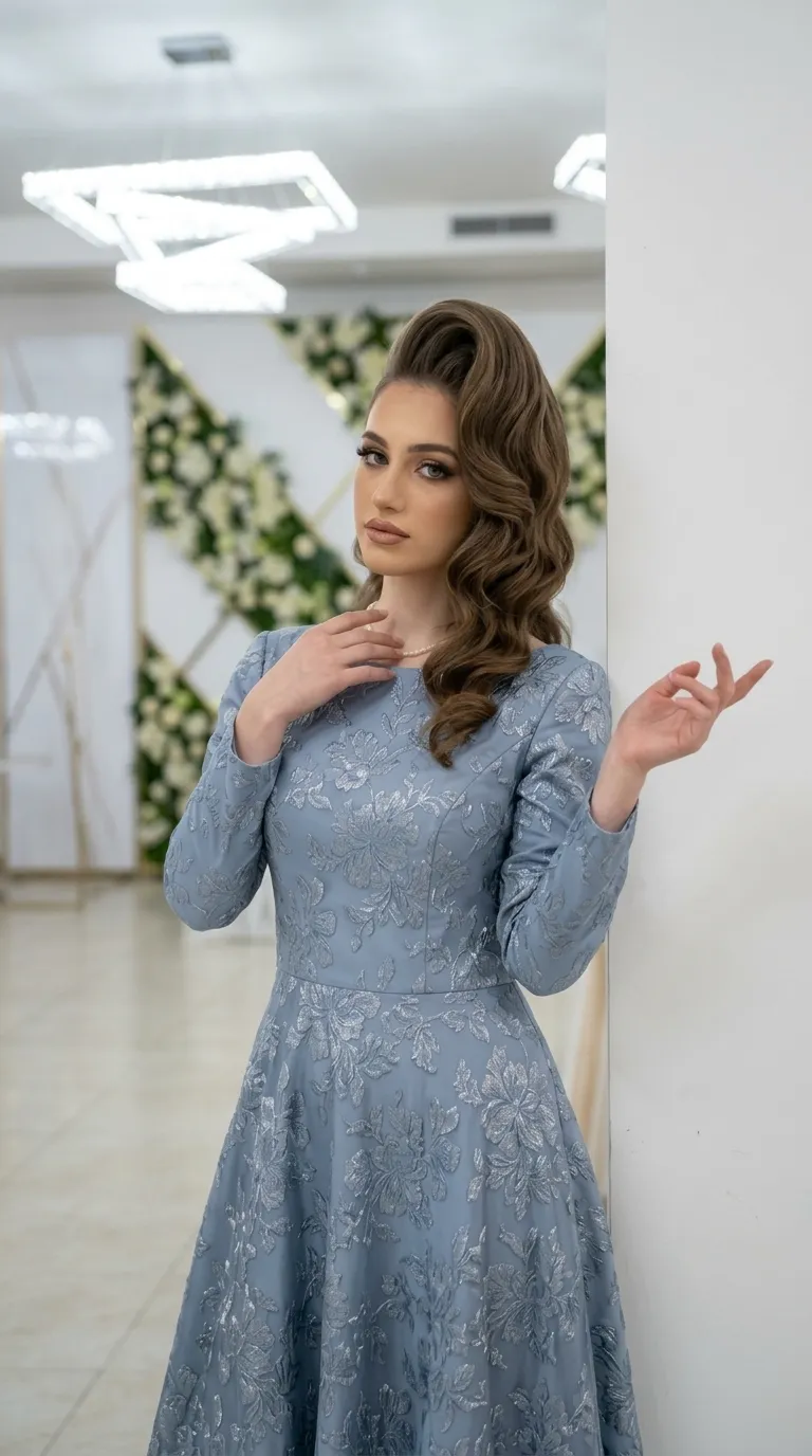שמלת ערב בגוון כחול מאובק העשויה מבד עשיר עם רקמת פרחים כסופה ובולטת לאורך כל פני השטח. השמלה מעוצבת בגזרת A-line עם קו מותן מודגש, וכוללת שרוולים ארוכים וצמודים וצווארון עגול וצנוע.
